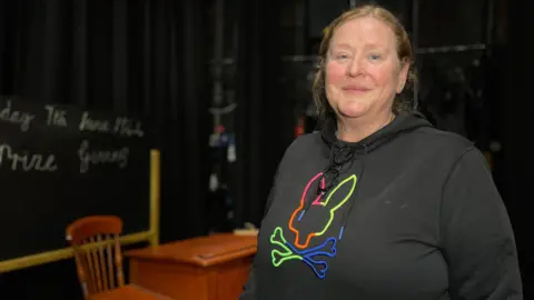 Maria smiles at the camera as she wears a black hoodie she has blonde hair tied into a ponytail. Behind her is a desk and a blackboard.