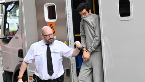 A man is escorted from a police van. He is handcuffed to a prison officer who has a white shirt on with a black tie. The prisoner is wearing grey tracksuit bottoms and a grey sweatshirt. He is covering his mouth with the grey sweatshirt and he has black hair.