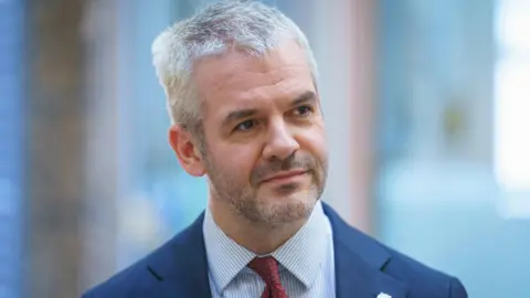 A man with white hair and a light beard turns his head slightly. He is dressed in a navy suit and maroon tie.