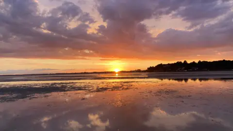 Ilovethebeach MONDAY - The sun glows orange on the horizon illuminating the sky and clouds in stunning orange colours and reflecting in the water of Poole Harbour. The still water reflects the sky and white clouds. In the distance on the far side of the harbour you can see houses nestled in the trees on the shore. 