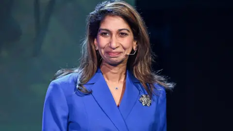 Suella Braverman is standing in front of a backdrop and in front of a black podium. She has brown hair and is wearing a blue suit jacket with black buttons. She has a large silver brooch on the jacket and a nude coloured microphone wire near her mouth