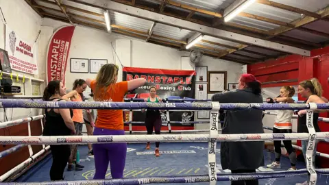 Nine women in a boxing ring. The floor is blue. The women are wearing sports clothes, including leggings and tank tops. The roof is slanted. The left and back wall is white and the right wall is red.