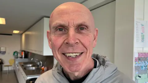 Terry Doyle, a bald gentleman dressed in a grey hooded jacket, smiles at the camera. He is standing in a kitchen area.