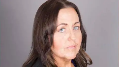Joanne Monk is pictured standing to the side with her head facing the camera. She has shoulder length brown hair.