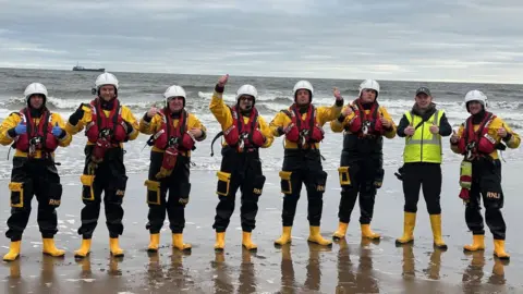 Alan Hughes. Ocho voluntarios del bote salvavidas, vestidos de rojo y amarillo, forman una fila y sonríen frente al mar. Todos levantan los pulgares hacia la cámara. Se ve un barco en el horizonte, detrás de ellos.