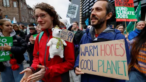 Climate change protesters take part in a march through Edinburgh in support of a global day of action against fossil fuels in September 2023. In the foreground is a woman in a red jacket with a toy polar bear and a sign saying "Get on with it" and a man in a blue jacket carrying a sign which says "warm homes cool planet". There are people holding placards in the background.