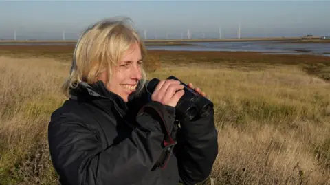 John Fairhall/BBC A blonde woman dressed in a black coat looks out to sea with her binoculars with wind farms in the background 