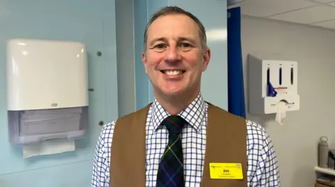 Ellen Knight/BBC Lacy-Colson photographed looking into the camera and smiling widely. He is wearing a white collared shirt with blue checks, a dark green tartan tie with red, yellow, and blue threads in it, and a light brown waistcoat. He has short brown hair, and on his left lapel he is wearing a yellow NHS name badge that reads 'Jon' in black letters. He is stood inside one of the clinic rooms - to his left is a glove dispenser, and to his right is a paper towel dispenser. The walls of the room are grey and light blue.