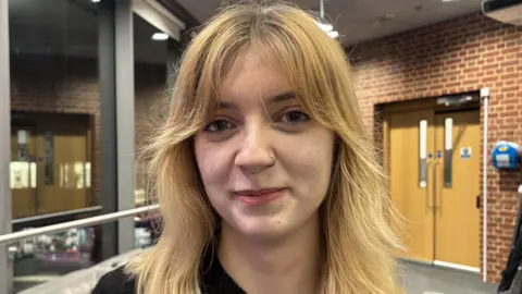 Grace Murray, standing in a room, looking straight at the camera, with long blonde hair and a nose ring on her left side. She is smiling. The building has a door to the right and red bricks. She is wearing a black top. 