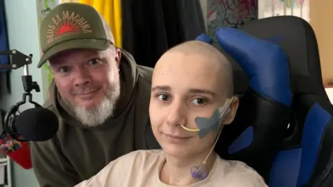 Family The boy from the above photos sitting in a gaming chair with a tube coming out of his nose. A man with a grey beard and a dark green baseball cap and hoodie is crouching down next to him. They are smiling at the camera.