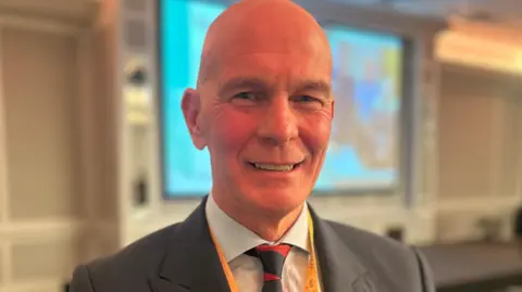 Mark Cox smiles at the conference, he is bald and wears a dark suit jacket, white shirt and red and black tie.
