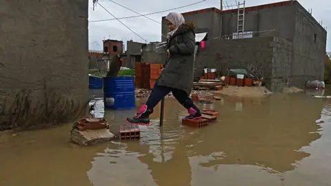AFP via Getty Images A woman wearing a headscarf walks through a flooded street in the Sidi Amor neighbourhood of Ariana district, near Tunis, with water covering the road around her