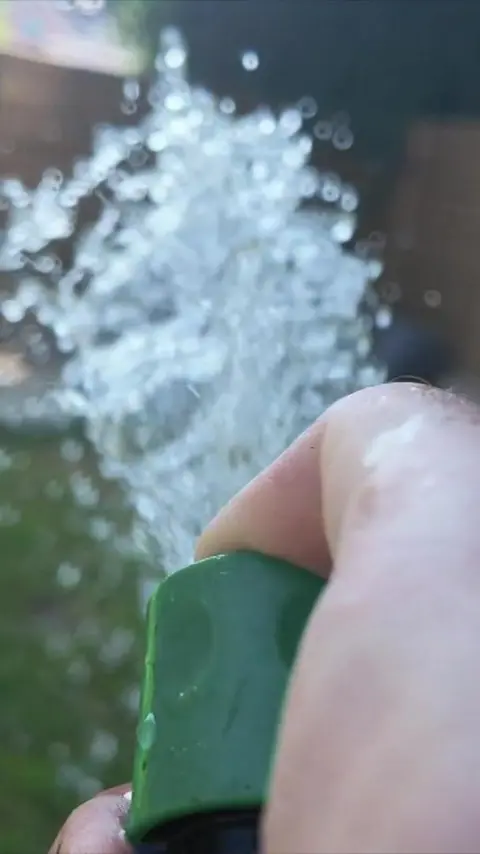 Water spraying out of a green hosepipe. A hand is holding the end of the hosepipe. 