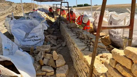 Construction workers wearing orange hi vis jackets and white construction helmets stand under canopies on a hot day as they work on building a Cotswold dry stone wall on a large construction site. They are surrounded by slabs of stone and large white construction bags.