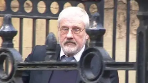 BBC Stephen Cox, wearing a buttoned-up shirt and tie, but with the top button undone, and a purple jumper and dark jacket, walking out of Reading Crown Court, with the bars of railings in front of him as he walks down a ramp outside the building