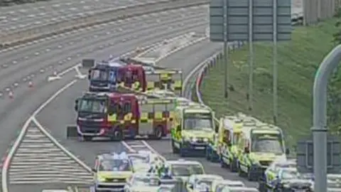 National Highways A grainy CC screenshot showing two fire engines and a several ambulances and a police car lined up on a motorway slip road.