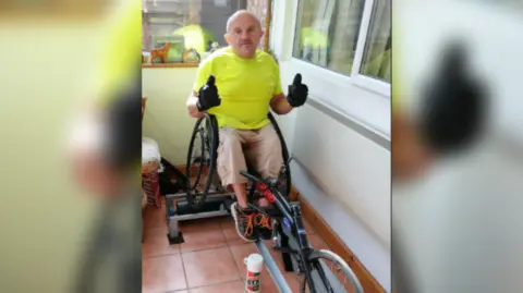 Les Hampton in his sports wheelchair in his conservatory at home, wearing a bright yellow t shirt and sand colour three quarter length cargo trousers. He has trainers on  and black gloves, with both hands in a thumbs up sign to the camera. 