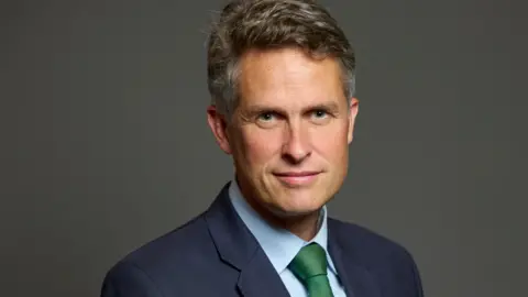 Sir Gavin Williamson MP is wearing a navy suit and green tie and is looks into the camera, in front of a dark grey screen.