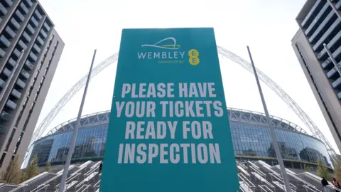A general view of the outside of Wembley Stadium with a blue sign stating 'Please have your tickets ready' in white lettering next to EE branding