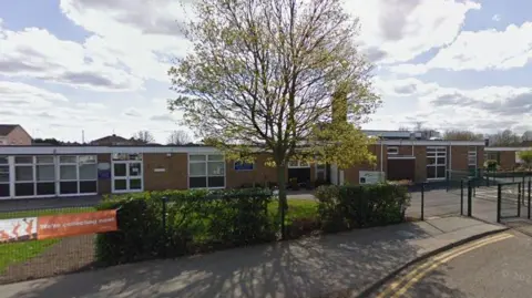 Exterior of a one-storey brick building with glass windows. There is some green space and a tree in front of the entrance. 