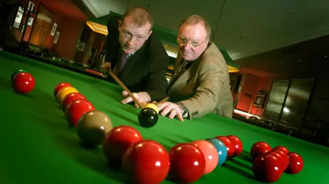 Steve Davies and Dennis Taylor pose at a snooker table, both appear to be aiming for the black ball. There are a number of snooker balls arranged in the front of the image.