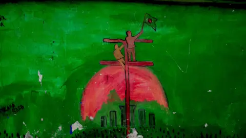 Mural showing outline of two people standing on a structure, waving a flat, on a background showing a sunrise in the colours of the Bangladeshi flag