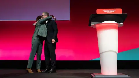 Getty Images Eluned Morgan hug on stage at the Welsh Labour conference. Eluned wears a grey suit and wearing a pink shirt, while Sir Keir wears a dark suit, white shirt, glasses and a dark red tie