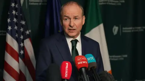 PA Media Micheál Martin stands in front of three microphones from Newstalk, Virgin Media news and RTÉ and is mid-speech. He is wearing a navy suit jacket, a green tie and a white collared shirt.