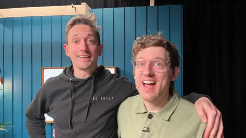 John Robins wore a black hoodi smiling with her arm around Ellis James, wearing green shirts and glasses, which smiles. You can see the blue panel walls of the podcast recording studio behind them