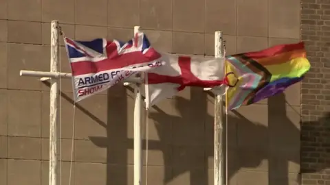 Three flags are fluttering in the wind outside a council building. They are an armed forces union flag, an England flag and a Progress Pride Flag. They are all flying from white poles.