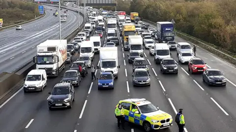 Six lanes of traffic on motorway have all been stopped with a police car parked in the centre. The cars are facing the camera and the image has been taken from  above the M25.