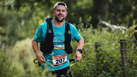 Sam Bagshaw A man in a blue t shirt running with a backpack on and wearing a cap, he has a number on which is 207.