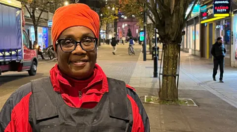 Holly Nichols/BBC Rose Forde is standing on a street at night, wearing a black stab-proof vest over a bright red jacket. The vest has yellow text that reads “STREETMOTHERS”. The street is lined with shops, including a brightly lit convenience store on the right and other businesses further down.