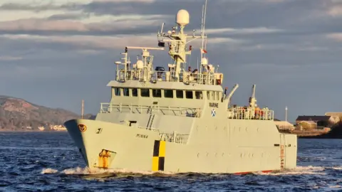Christopher Brindle A grey painted ship, Minna, photographed as it sails at a slight angle towards the camera