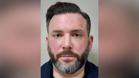 A police mugshot of a man with black hair and a black beard with some grey hairs. He is facing the camera and is wearing a dark blue top.