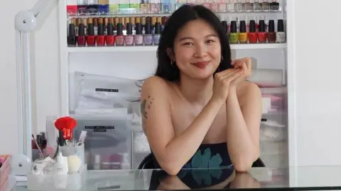 Nail artist Jess Young Nail artist Jess Young sits behind a table and smiles at the camera. On the table there are nail tools and behind her are nail polish bottles in a range of colours. 
