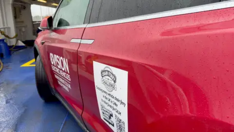 Image shows a red Tesla which was used to help the team complete the challenge. It's on a ferry from the Isle of Wight to Lymington. On the side, is it a sticker which says 'Chain ferry challenge' and has a QR code for people to donate.