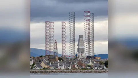 Quintin Lake Some large metal rigs towering above some houses on the coast at Ross & Cromarty, Scotland.