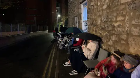 Bella Barnes A row of people sitting on chairs along a pavement with the wall of the letting agent building behind them. A lot of the people are wearing coats and hats, and some have umbrellas up.