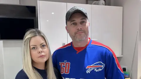 Kate Bradbrook/BBC Woman on left with long blonde hair stands in a kitchen next to man in a baseball cap and blue sports shirt.