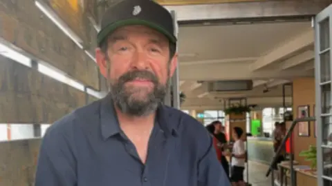 Andrew Small Andrew Small, who has a black beard and is wearing a black baseball cap and dark blue shirt, smiles into the camera.