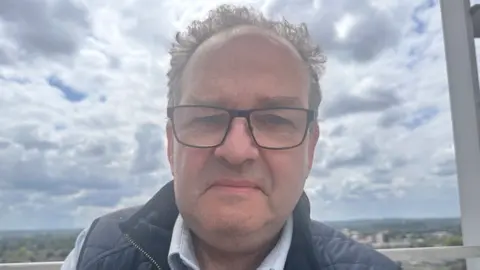 Mike Swift, who has a light blue shirt and a dark blue gilet on. He has dark glasses and wavy, dark hair