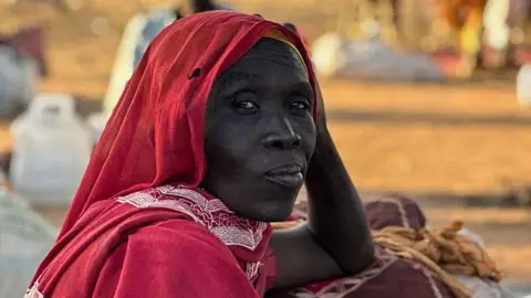 A displaced Sudanese woman who fled el-Fasher after the city fell to the Rapid Support Forces is wearing a red garment. She is sitting on the ground among bags, blankets and metal containers in an outdoor setting, with other people and belongings visible in the background.