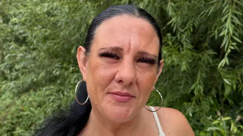 Sandra McCluskey with long dark hair stands outdoors beside a wooden fence, with dense green trees and foliage in the background. She is wearing a light-coloured sleeveless top and large hoop earrings, and has a visible tattoo on the upper arm. The photograph is a close-up portrait taken in natural daylight, with the surrounding greenery filling most of the frame.
