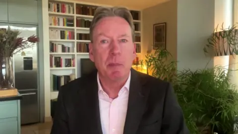 BBC Frank Gardner sitting in his home being interviewed over Zoom. He is wearing a very light pink shirt and dark jacket, with a bookshelf and plants visible behind him.