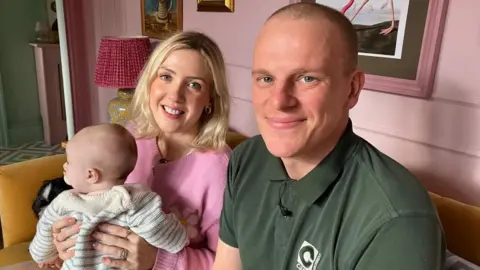 Jessica and Rick Sowerby sit on a sofa with their baby son on Mrs Sowerby's knee. Mrs Sowerby has bobbed blonde hair and is wearing a pink jumper. Mr Sowerby wears a green polo shirt. Both are smiling. Their son wears a grey and white striped jumper. The couple's dog Merla can just be seen on the sofa behind them.