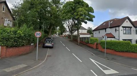Woodgate Avenue with tall leafy trees and detached houses on either side. A road sign showing a 20mph speed limit stands on the corner.