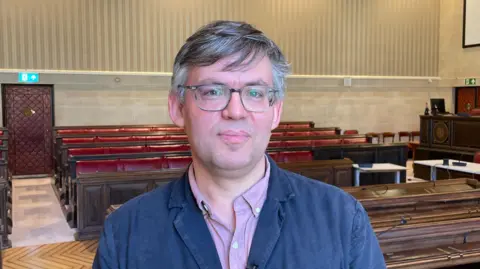 Barry Parson smiles at the camera. He is standing in a council chambers, with wooden pews seen behind him. He is wearing a dark blue jacket and a light purple shirt. He is wearing glasses.