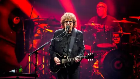 A man with long curly brown hair in dark clothing, holding a black guitar in front of a band lit with red lights