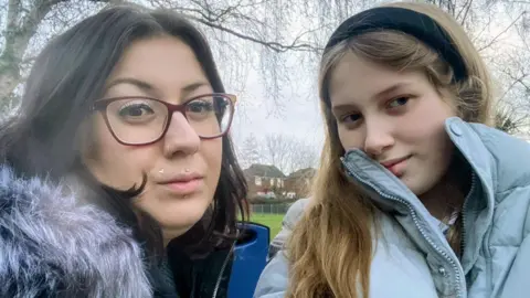 Selfie-style picture of Rhiannon Van Der Zanden and her daughter Alexis. Rhiannon has black hair and glasses and wears a coat with a fur collar. Alexis has long blonde hair worn with a black alice band. Her coat is light blue. 